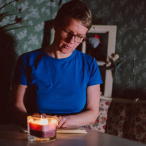 Frau sitzt am Tisch bei Kerzenlicht, schaut auf das Papier vor sich und schreibt etwas mit einem Stift