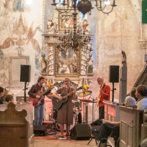 Eine Band steht vor dem Altar einer Kirche und spielt Musik. Im Anschnitt ist Publikum zu sehen auf den Bänken davor.