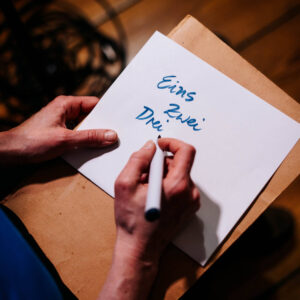 Die rechte Hand hält einen Stift, die linke hält ein Blatt Papier auf dem geschrieben steht: Eins Zwei Drei