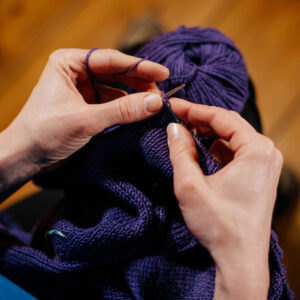 Zwei Hände die Stricknadeln halten, darunter eine Strickarbeit in Violett