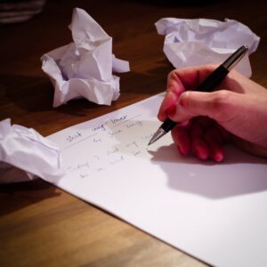 Eine Hand, die einen Stift hält setzt zum Schreiben an auf einem Blatt Papier. Drumerhum sieht man drei zerknüllte Papierblätter liegen.